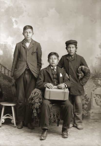 Studio portrait in front of a painted backdrop of a Ho-Chunk boy posing sitting, and two boys standing. The young man sitting is holding a a package wrapped in paper, most likely containing his belongings for boarding school. He is dressed in a suit and necktie. Standing on the left, John Stacy Jr. (HaGaKah), is wearing a suit coat and is holding a cap in his right hand. The unidentified boy standing on the right is wearing a heavy coat and cap and is holding a folded shawl under his left arm.