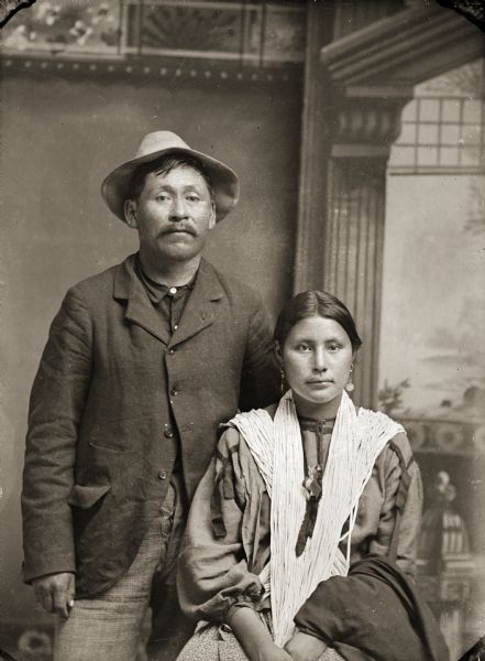 Studio portrait of Nebraska Winnebago John Clay (WaCheDaKaw) posing standing in front of a painted backdrop on the left. He is wearing a suit and hat. Grace Decorra Clay Whitegull (BeekSkaWinKah), Ho-Chunk, is posing sitting next to him on the right, and is wearing a finger braided shawl and earrings.