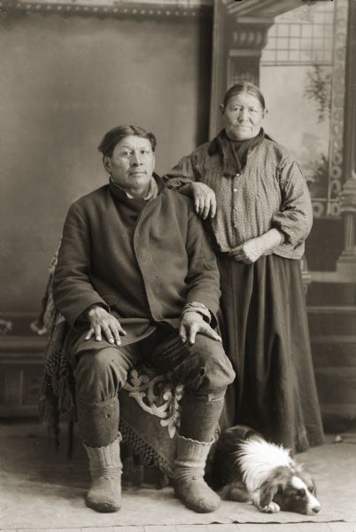 Full-length studio portrait of a Ho-Chunk couple, John Buffalohead and wife Mary Snowball Decorra Buffalohead, with their dog lying at their feet. Clans kept an inventory of clan names of special dogs. In the background is a painted backdrop. John Buffalohead (ChayPaKah) was born in 1862 and died July 9th, 1914. (Son of [JumpBroHunWinKah] Mary Pettibone and 1/2 brother by mother to Peter Rockman.) John is the father to: Frank Buffalohead (WahChoZhoNoZhooKah), Kate Buffalohead (ENooKah) and Abel Buffalohead (MaNaKasRayHeKah). Mary Snowball, Decorra, Buffalohead (PayDokSeKah [Edge of brow is yellow]) was born in 1863 and died on November 20th, 1940. (Daughter of [WauSooMonEKah] Snow Ball and [WaConChaWinKah]) (Maternal granddaughter of Iron Walker and Green Feather Woman). Mary Snowball Decorra Buffalohead was married to Tall Decorra (HayNunkSeRatehKah) previously.