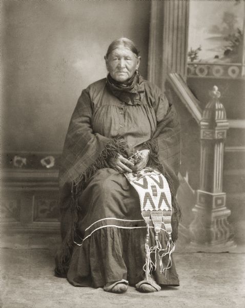 Full-length studio portrait of an elderly Ho-Chunk woman, Mary Snowball Decorra Buffalohead (PayDokSeKah [Edge of brow is yellow]), sitting and holding a porcupine quill-work bag. The pipe bag is not of Ho-Chunk origin. In the background is a painted backdrop. She was born in 1863 and died November 20th, 1940. She was the wife of John Buffalo Head (ChayPayKay) and the mother of five children: Grace, John, Stella, Frank and Carrie. (Daughter of Snow Ball [WauSooMonEKah] and [WaConChaWinKah]) (Maternal granddaughter of Iron Walker and Green Feather Woman).