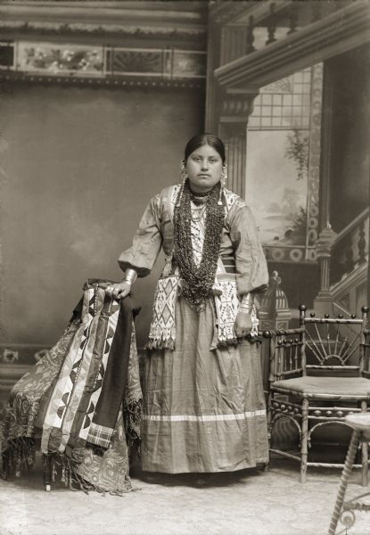 Full-length studio portrait in front of a painted backdrop of a Ho-Chunk woman, Ida Lizzie Decorra Blowsnake (Real Wampum Woman) (WooRooShiekESkaeWinKah), wife of George Lowe, posing standing and wearing a large necklace made of beads that partially covers the bandolier bags crossed over her shoulders. She is wearing German silver rings and bracelets on her fingers and wrists. To her left, a Ho-Chunk applique blanket is draped on a chair.