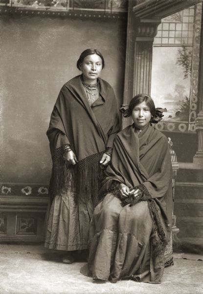 Full-length studio portrait in front of a painted backdrop of two Ho-Chunk women, Jennie Youngthunder Decorah (PetHatChaCooWinKah) standing on the left, and Annie Thunderking Lowe Lincoln (WaNukScotchEWinKah) sitting on the right. Both are wrapped in fringed shawls.