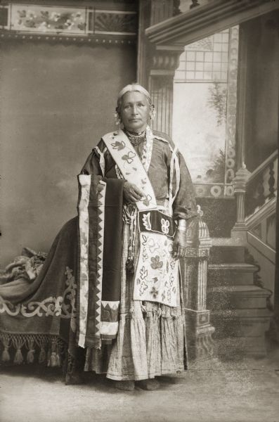 Full-length tudio portrait of Ho-Chunk elder, Mary Thunderchief Snowball (WaSayMaKaWinKah), posing standing in front of a painted backdrop. She is holding a silk applique blanket on her arm and has a floral applique beaded bandolier bag hanging across her right shoulder. She is wearing short bead necklaces, file bracelets, and short coin earrings.