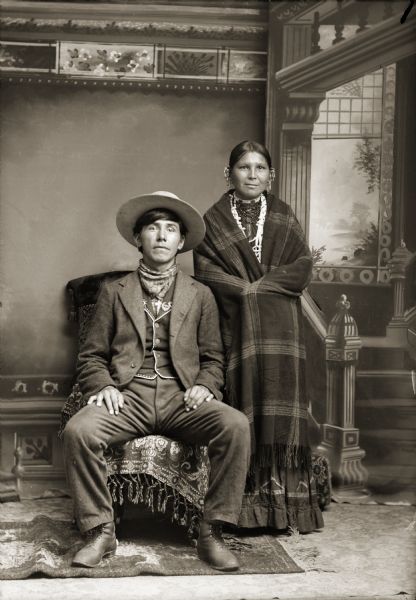 Full-length studio portrait of Amos Wallace (WeeKah), a Ho-Chunk-Menominee and his Ho-Chunk wife, Annie Bearchief Wallace Bearheart Redbird (MaHaNaKayWinKah) posing in front of a painted backdrop. He is sitting in a chair and is wearing a suit jacket, vest, trousers, and hat. She is standing and is wearing a plaid shawl, beaded necklaces, and ear bobs. It was not uncommon for the Ho-Chunk to marry outside the tribe. In fact, at certain times in their history it was necessary in order to increase the population, which was sometimes reduced by disease and other catastrophes. Annie Bearchief is also the mother of Joe Green according to Jackson County Historical Society notes.