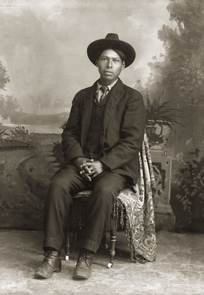 Full-length studio portrait in front of a painted backdrop of Charlie Youngthunder (MahAhQuaKah), a Ho-Chunk man. He is posing sitting with his hands loosely clasped in his lap, and is wearing a suit, necktie, and hat.