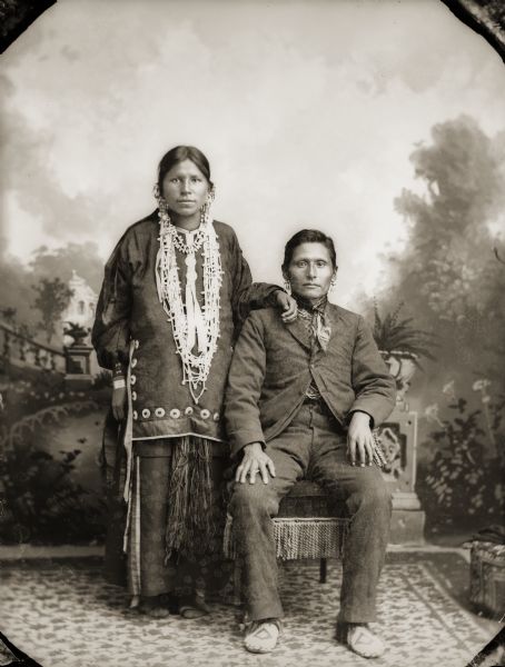 Full-length studio portrait in front of a painted backdrop of a Ho-Chunk couple, Annie Waukon (HumpMeNuKah) and Luke (Duke) Snowball (NySaGaShiskKah). Annie Waukon, left, is wearing a traditional blouse, several necklaces, earrings, file bracelets, moccasins, and a finger woven sash. Luke Snowball is posing sitting on a chair next to her. He is wearing a suit, bandana, earrings, and beaded moccasins.