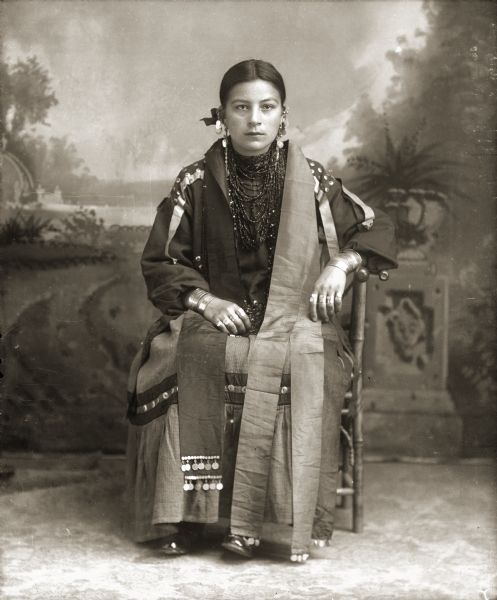Studio portrait of a Ho-Chunk woman, Alice Mary Blackdeer Hopkinah (NautchGePinWinKah), posing sitting in front of a painted backdrop. She is wearing a woman's dress (hinukwaje) with long strips of ribbon over her shoulders and silver coins attached to the bottom. She is also wearing several necklaces, earrings, rings, file bracelets, and coin-trimmed stoles.