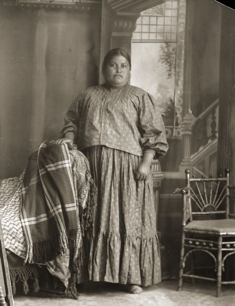 Full-length studio portrait in front of a painted backdrop of a Ho-Chunk woman, Ida Lizzie Decorra Blowsnake (Real Wampum Woman) (WooRooShiekESkaeWinKah), wearing a plain dress (hinukwuaje) and moccasins. She is posing standing with her hand on a chair covered by a plaid wool shawl. 	
