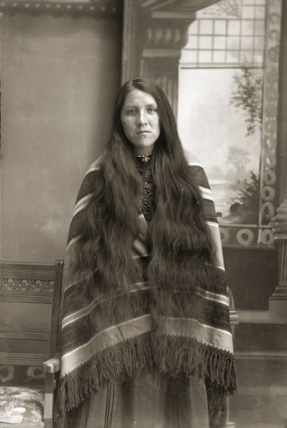 Three-quarter length studio portrait of a Ho-Chunk woman, Mamie (Minnie) Bearchief Funmaker (HoHumpCheKaRaWinKah) posing standing near a chair in front of a painted backdrop. She is wearing a dress with a striped wool shawl, and her hair is loose over her shoulders. She is wearing a necklace and brooch on her collar. According to the Indian Census Roll taken at Tomah Indian School reservation in 1930, Mamie Bearchief was the wife of Ed Funmaker and mother of Samuel and Verna Funmaker.