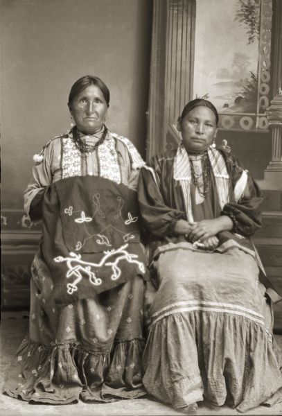 Studio portrait of two Ho-Chunk women posing sitting in front of a painted backdrop. Both woman are wearing dresses, earrings, and necklaces. The woman on the left, holding the unfinished beaded blanket  over her lap, is Mamie (Minnie) Bearchief Funmaker (HoHumpCheKaRaWinKah), wife of Ed Funmaker. She is also wearing a beaded shirt typically worn by men. On the right is an unidentified woman.