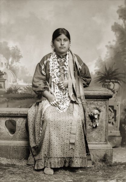 Full-length studio portrait of a Ho-Chunk woman, Ida Lizzie Decorra Blowsnake (Real Wampum Woman) (WooRooShiekESkaeWinKah) posing sitting on a prop stone wall in front of a painted backdrop in Van Schaik's studio. She is wearing a traditional woman's dress (hinukwaje) made from brightly painted fabric. Solid-color cloth was usually used to make Ho-Chunk women's dresses.