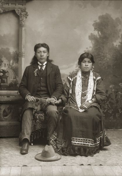Full-length studio portrait in front of a painted backdrop and prop stone wall of a Ho-Chunk couple, Henry Greencrow (CooNooZeeKah), left, and wife Belle Hall Greencrow (AhHooSkaWinKah), right. They are posing sitting and holding hands. He is wearing a suit jacket, vest, tie, and trousers, and his hat is on the floor in front of him. She is wearing a dress, earrings, rings, bracelets, and long strands of beads. According to the U.S. Indian Census Roll of 1920, Belle Hall Greencrow was the mother of Paul, Fannie, Annie, Nettie, Mary and Henry K. Greencrow.