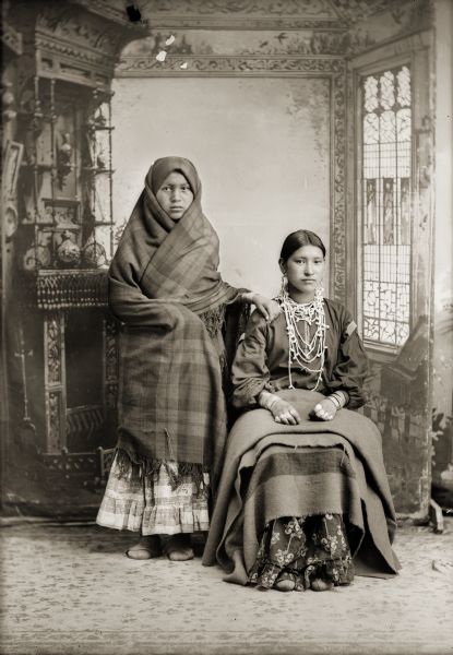 Full-length studio portrait in front of a painted backdrop of two Ho-Chunk women. The woman on the left, possibly Miss Starr, is standing and is wrapped in a Racine Woolen Mills shawl with her hand on the other woman's shoulder. The woman sitting on the right is wearing German silver earrings, bracelets, and rings and has a Hudson's Bay trade blanket on her lap. She is identified as Mrs. Whitegull, Frank Johnson's aunt.