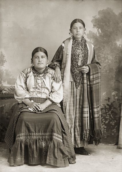 Full-length studio portrait in front of a painted backdrop of two young Ho-Chunk women, Ida Lizzie Decorra Blowsnake (Real Wampum Woman) (Woo Roo Shiek E Skae Win Kah), sitting, with half-sister, Johanna Puss Otter Greendeer Tebo (Wa Ga Cha Me Nuk Kah). Both women are wearing regalia including several necklaces and earrings. Johanna Puss Otter is wearing a plaid print skirt and holding a Racine Woolen Mills shawl, while standing with her hand on Ida Lizzie's shoulder.