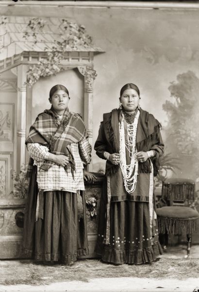 Studio portrait of two Ho-Chunk women, Minnie Pigeon Whiteotter Blowsnake Davis (AhHooKeShelNWinKah), left, and Emma Blowsnake Battise Good bear Walking Priest Littlejohn Mike (ChakShipEWinKah), right, posing standing in front of a painted backdrop and low prop stone wall. Minnie Pigeon is wearing a plaid shawl over her shoulders, and the woman on the right is wearing several necklaces earrings, rings, file bracelets, and fringed mantelet. Emma Blowsnake is probably the wife of John Mike, and Minnie Pigeon is probably the wife of John Davis according to Jackson County Historical Society notes.