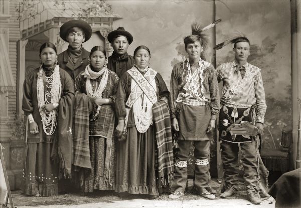 Studio portrait of group in front of a painted porch backdrop, typical of many Van Schaick's photographs. The three Ho-Chunk women are wearing several necklaces, earrings, and file bracelets and holding plaid shawls. Two of the men are wearing bandoleers and feathers, the two other Ho-Chunk men are standing and wearing hats. Identified from left to right as Emma Blowsnake Batitise Goodbear Walking Priest Littlejohn Mike (ChakShipEWinKah), Amos Wallace (WeeKah), NoKetteKyWinKah, Will Stohegah Carriman (WonkShiekStoHeKah), Mary Goodvillage Hindsley (ChoWasSkaWinKah), William Hindsley (Hensley) (CooNooChoNeeNikKah), and Edward Winneshiek (NaZhooMonEKah).