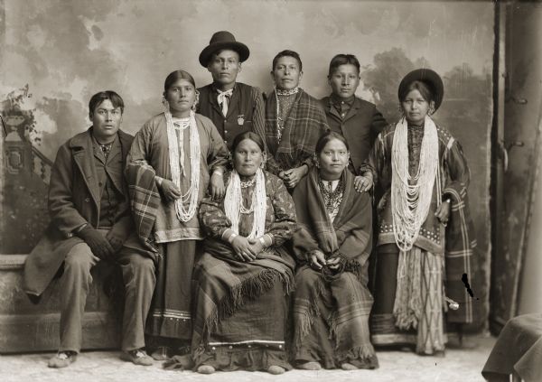 Studio portrait in front of a painted backdrop of four Ho-Chunk men and four Ho-Chunk women posing sitting and standing. From left to right in the back are Charles George Whiteotter (HoonkHaTaKah), John Lewis (John Johnson) (NoCheKeKah) and Will Stohegah Carriman (WonkShiekStoHeGah). From left to right in front are George Blackhawk (WonkShiekChoNeeKah), Minnie Pigeon Whiteotter Blowsnake Davis (AhHooKeShelNWinKah), Mary Mollie Prophet Thunder (HoWaChoNeWinKah) and an unidentified woman. Standing on the right is Charles Low Cloud's sister Mary L. Decorah (HoChumLaWinKah). George Blackhawk is dressed in traditional Ho-Chunk men's moccasins that do not have a top flap. Many of the women are wearing several necklaces, earrings, and shawls, while most of the men are wearing contemporary suits and clothing.