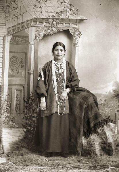 Full-length studio portrait of a Ho-Chunk woman, Mary Alice Johnson Decorra Brown (ENooKah), posing standing in front of a painted backdrop. SHe is wearing beads, earrings, and file bracelets. Probably the sister of John Johnson.