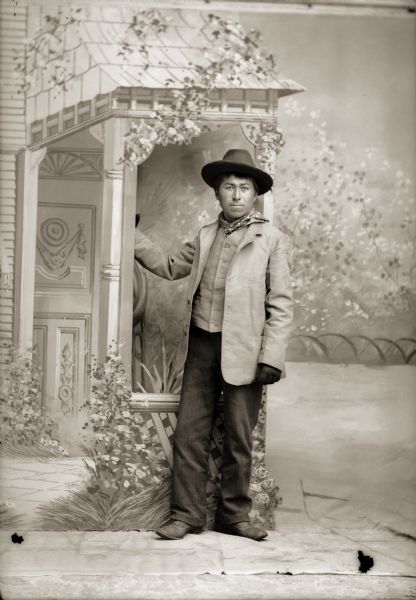 Full-length studio portrait of a young Ho-Chunk man, Charlie Smith (John Beaver) (WySkaInKah), standing and wearing a suit and hat. He is posing near a prop front porch in front of a painted backdrop.