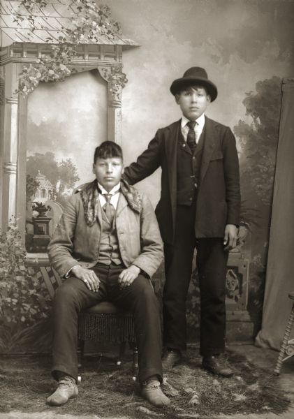 Full-length studio portrait of two young Ho-Chunk men posing in front of a painted backdrop. They are both wearing suits and ties. The unidentified man sitting on the left is wearing a piece of fabric around his shoulders, and Ho-Chunk moccasins with his suit and tie. The man standing on the right, Frank Big Soldier (KeKahUnGah), is wearing a hat and has a hand around the other man's shoulders.