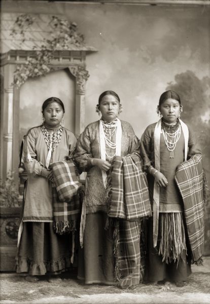 Full-length studio portrait of three young Ho-Chunk women. They are, from left to right: Grace Decorra Twocrow Winneshiek Massey (MauHeHutTaWinKah), Florence Littlesoldier Wallace Thunder (MauKonNeeWinKah), and Alice Standstraight Blowsnake Littlewolf (HeChoWinKah), posing in front of a painted backdrop. They are standing and wearing several necklaces, earrings, and file bracelets, and are holding a Racine Woolen Mills shawls over their left arms.