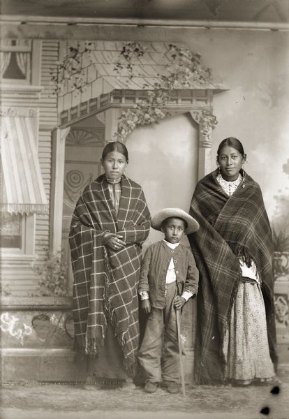 Studio portrait in front of a painted backdrop and prop stone wall of a Ho-Chunk boy and two young Ho-Chunk women. The boy is standing and wearing a hat and is holding a stick. The two young Ho-Chunk women are standing and wearing earrings, and are wrapped in plaid shawls/blankets. One of the women may be the wife of George Otter.