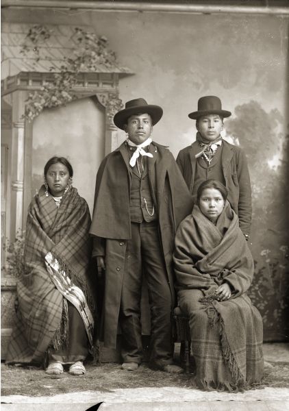 Studio portrait in front of a painted backdrop of two Ho-Chunk women posing sitting in front of two standing Ho-Chunk men. The women are wrapped in shawls, and the woman on the right has several earrings. The men are wearing hats, suits, and bandanas, and the man on the left is also wearing a Spanish-American War overcoat. From left to right, Maude Climer Winneshiek (AhHooKeShinNeWinKah), David Bow Littlesoldier (MauJchayMauNeeKah), an unidentified man, and Kate Whitespirit-Dickson Thunder Decorra Baptiste Seymour (NeChooKoontchRaWinKah) in front. Maude is wearing a silk applique blanket covered by a Racine Woolen Mills shawl and traditional Ho-Chunk moccasins.