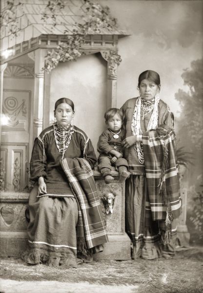 Studio portrait in front of a painted backdrop of two Ho-Chunk women and a boy. Lucy Decorah (AhHoRaPaNeeWinKah) is sitting on a prop stone wall on the left, and Belle Hall Greencrow (AhHooSkaWinKah) is posing standing on the right holding a small Ho-Chunk boy on the prop stone wall column in between them. Both women are wearing several necklaces, earrings, and file bracelets, and have plaid shawl/blankets draped over their right arms. The boy is wearing a necklace and medallion.