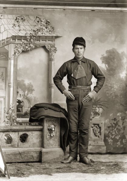 Studio full-length portrait in front of Van Schaick's gazebo backdrop of a Ho-Chunk man, wearing period clothes, standing next to a prop stone wall draped with an overcoat. He has his hands on his hips and is wearing a woven fringed sash, dark shirt, necktie, and hat.