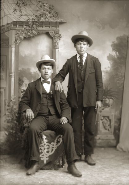 Studio portrait in front of a painted backdrop of two young Ho-Chunk men, Frank Bigsoldier (KeKahUnGah), sitting on the left, and a man possibly identified as Julius Whitedog (ChakShepWasKaHeKah) standing on the right. Both men are wearing hats, ties, vests, and slacks.