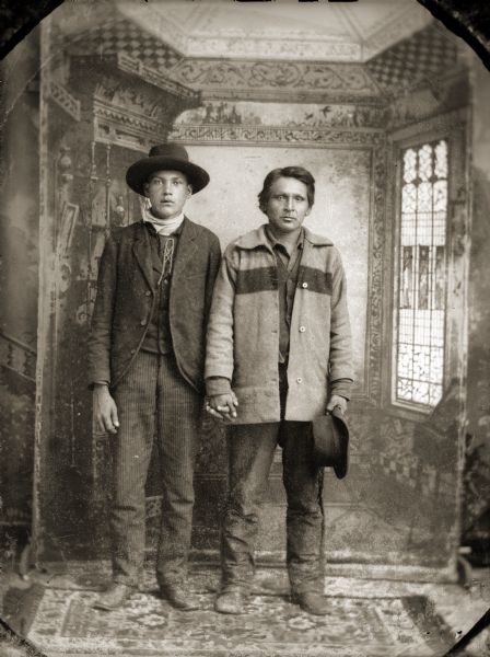 Studio portrait of two Ho-Chunk men posing standing in front of a painted backdrop and holding hands. The young man on the left is wearing a suit, bandana, and hat, and the man on the right is wearing a Hudson Bay wool sweater/coat and holding a hat in his left hand. The man on the right is identified as Four Cloud according to the Jackson County Historical Society of Wisconsin. Both are believed to be father and son.