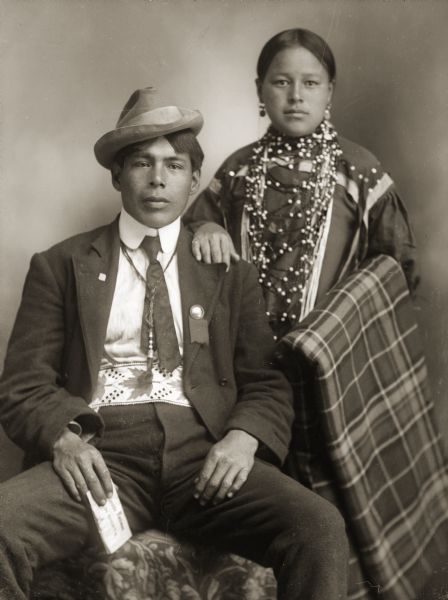 Studio portrait in front of a painted backdrop of a Ho-Chunk couple, Albert A. Johnson (HunkChoKah) and Annie Bessie Arthur Johnson Standingwater (WeHunKah). Annie Bessie is holding a blanket over her arm and is wearing earrings and a necklace. Albert Johnson is sitting and is wearing a hat. They are both wearing a mix of traditional and contemporary clothing.