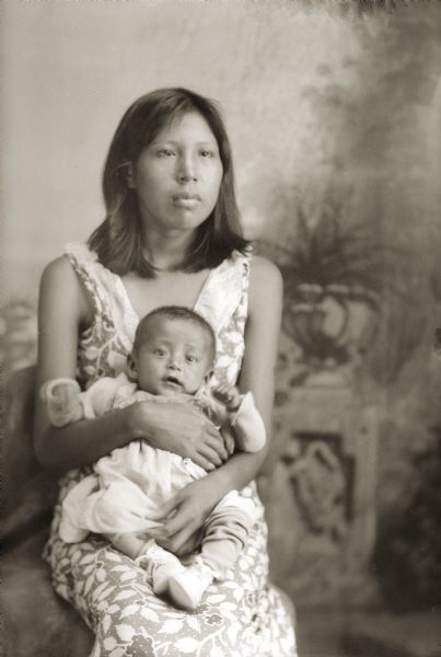 Studio portrait of a Ho-Chunk woman, Marie Big Hawk Whitewater, holding her baby Clyde White in her lap in front of a painted background. She has shoulder length hair and is wearing a sleeveless dress.