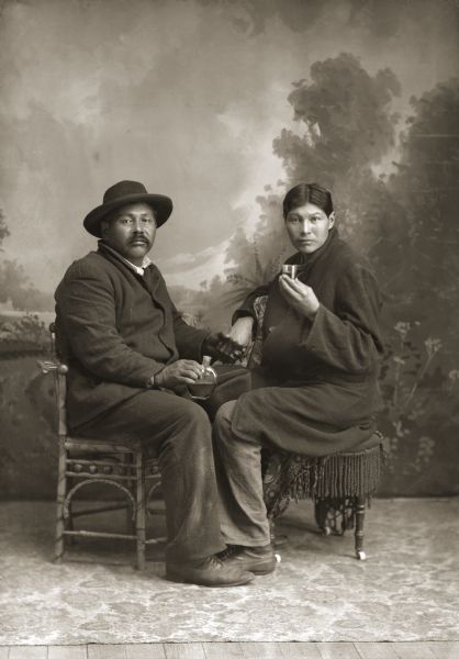 Full-length studio portrait of two Ho-Chunk men sitting at a small table. Louis Lookingglass (MaukHeKah), left, is holding a liquor flask and is wearing a hat. The other man (unknown) is holding a glass and wearing a long coat. In the background is a painted backdrop.