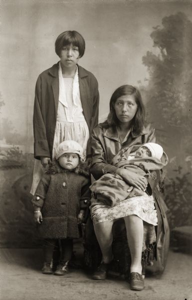 Studio portrait in front of a painted backdrop of a Ho-Chunk woman, Amanda Bighawk posing sitting holding her son Calvin Smith-Whitewater, and a Ho-Chunk woman, Marie Big Hawk, posing standing behind her daughter Betty Walker. They are all wearing contemporary clothing.