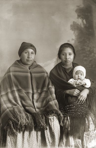 Studio portrait in front of a painted backdrop of two Ho-Chunk women and  and infant. On the left is Mae Mary Smith Whitewater (WaRaKaKaKah), with daughters Mary Whitewater Littlegeorge (HoUpSootchAWinKah), and Berdine Whitewater Tebo Littlejohn on her sister's lap. They are wrapped in five-row beaded fringe shawls. Mary White Littlegeorge is the wife of Yellow Bank. Mae Mary Smith Whitewater is the wife of Little George according to the notes of Jackson County Historical Society, Wisconsin.