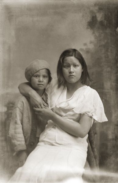 Waist-up studio portrait of two Ho-Chunk girls posing in front of a painted backdrop. The older girl, Viola Stacy, sitting on the right, is wearing a white dress, and has her arm around the shoulders of her younger sister, Doris (Dolly) Stacy, who is standing on the left wearing a beret. Close-up style photographs featuring contemporary dress were taken late in Van Schaick's career.