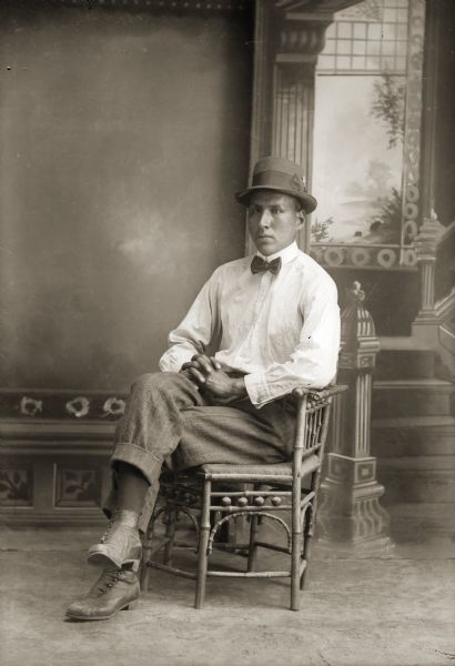 Studio full-length portrait in front of a painted backdrop of a Ho-Chunk man posing sitting in a chair with his legs crossed and hands clasped. He is wearing a light-colored shirt, a bow tie, and a hat.