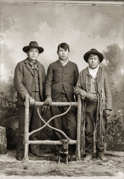 Studio portrait in front of a painted backdrop of three Ho-Chunk men posing standing behind a prop wooden fence. They are all wearing differently cut suit coats. The unidentified man on the left is wearing a hat and bandana, Will Blowsnake (One Who Stands and Strikes) (WoeGinNawGinKah) in the center is wearing a bandana, and Charlie Smith (John Beaver) (WySkaInKah) on the right is wearing a hat, loose bandana, and a finger woven sash. In front of the men is a small dog, whose head is blurred in motion, sitting in the center in front of the fence.