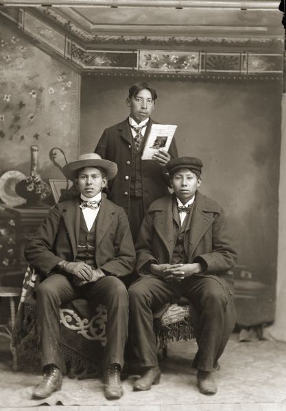 Studio portrait in front of a painted backdrop of two young Ho-Chunk men posing sitting in front of a man standing behind them. The two men sitting are wearing suits, bow ties, and hats, with the man on the right, David Davis W. Decorra (NeZhooLaChaHeKah), also wearing an overcoat. The man standing, Robert Sine Jr., is holding a magazine in his left hand, and is wearing a suit and necktie.