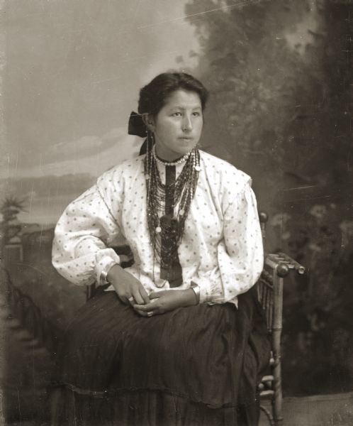 Studio portrait of a young Ho-Chunk woman, Minnie Sherman Prettyman-Bear Whitebear (AhHooSootchPaWinKah), posing sitting in front of a painted backdrop. She is wearing a contemporary light-colored printed blouse, a bow in her hair, and several necklaces, earrings, rings, and file bracelets.