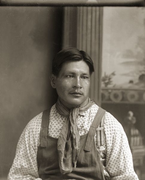 Waist-up studio portrait of a Ho-Chunk man, Henry (Little) Snake (HaRaChoMonEKah), sitting and wearing a scarf around his neck in front of a painted backdrop. He is wearing overalls, a bib, a contemporary work shirt, neckerchief and suspenders over his shirt. This is one of the few Van Schaick photographs where the subject is not looking directly at the camera.