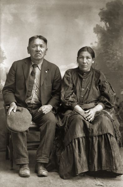 Full-length studio portrait of an elderly Ho-Chunk man and woman posing sitting in front of a painted backdrop. He is wearing a suit jacket, shirt with watch fob, tie, and trousers and is holding a hat. She is wearing a long dress. Identified as Charles Greencrow, a Nebraska Winnebago and his wife, Belle Blackhawk Monegar Cassiman Greencrow (AwHooSuchRayWinKah). She was the former wife of Arthur Cassiman, and sister of John Blackhawk according to Jackson County Historical Society notes.