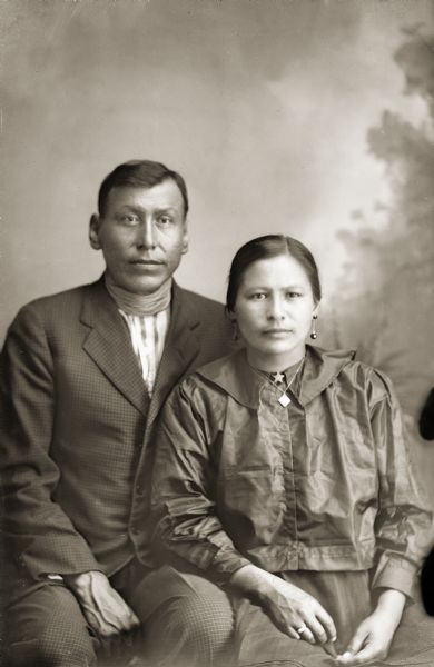 Three-quarter length studio portrait of a Nebraska Winnebago man and Ho-Chunk woman posing sitting and wearing modern dress in front of a painted backdrop. Identified as Oscar Raisewing, possibly of the Nebraska Thunder clan, with his wife, Louisa (Marie) Johnson Thunder Raisewing. They were probably members of the Native American Church or "peyote religion" according to Jackson County Historical Society notes.