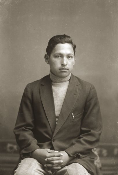 Three-quarter length studio portrait of a young Ho-Chunk man posing sitting in front of a painted backdrop. He is wearing a double-breasted jacket and turtleneck sweater. Possibly Ernst or Howard White Eagle, who was a follower of the "peyotism" or Native American Church according to the notes of Jackson County Historical Society, Wisconsin.