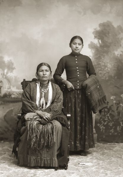 Studio portrait in front of a painted backdrop of two Ho-Chunk women. On the left, a woman is posing sitting and is wearing traditional clothing with several necklaces, earrings, and rings. To the right, a woman is posing standing and wearing a western-style blouse with a long row of buttons while holding a wool shawl decorated with German silver brooches over her left arm.