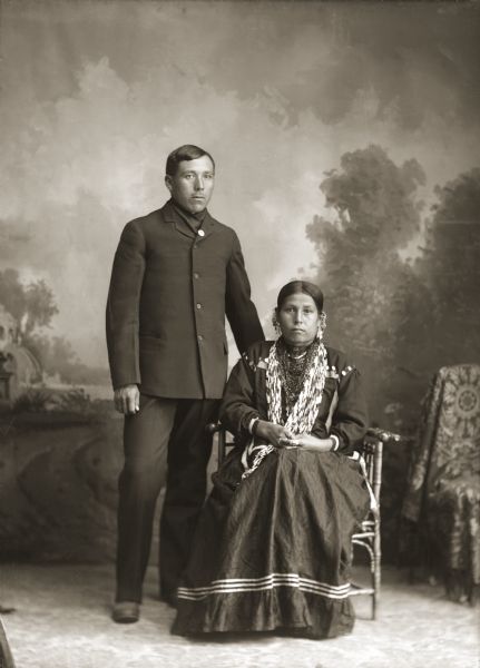 Full-length studio portrait in front of a painted backdrop of a Ho-Chunk couple in front of a painted backdrop. The man is posing standing on the left and is wearing a suit coat. The woman is posing sitting on the right and is wearing many necklaces, earrings, rings, and a decorated blouse. They are identified as Martin Green (Snake) (KeeMeeNunkKah) and Dora Monegar Wallace Green (Snake) (ChePinChayWinKah).