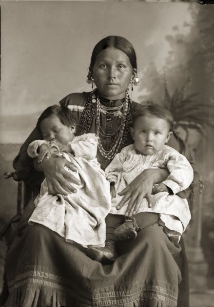 Studio portrait of Mountain Wolf Woman, also known as Stella Blowsnake Whitepine Stacy (HayAhChoWinKah), sitting and holding her two daughters, Josephine Whitepine Mike (AhHooGeNaWinKah), left, and Lena Whitepine Shegonee (HaCheDayWinKah). <i>Mountain Wolf Woman</i> is also the title of a book written about her by Nancy Lurie.