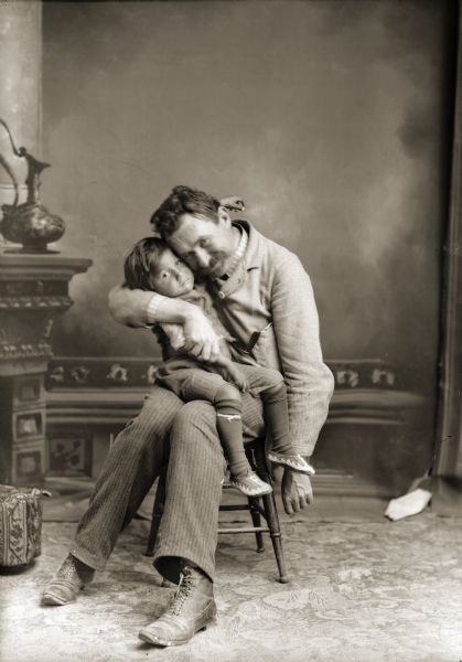 A studio portrait of Charles Van Schaick, sitting on a stool in front of a painted backdrop, holding and hugging a small boy on his lap.