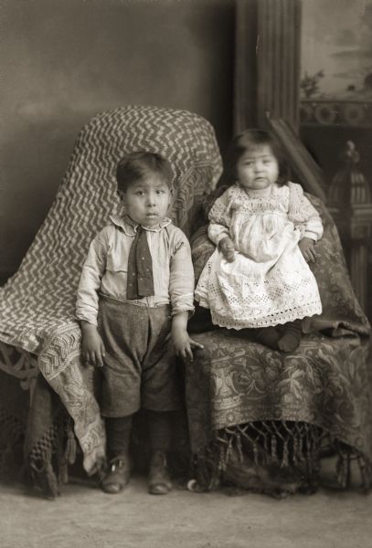 Full-length studio portrait in front of a painted backdrop of a Ho-Chunk boy standing and girl. The boy is wearing short trousers, shirt, and tie, and the girl is wearing a lace dress, with her face slightly blurred from movement, and sitting in a chair. Identified as Daniel Greengrass and Ruth Greengrass, probably the children of Irvin Blackdeer and Bertha Greengrass Blackdeer.